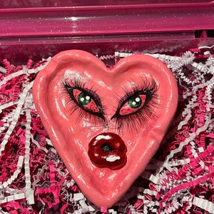 May include: A pink heart-shaped trinket dish with painted eyes and lips. The eyes have long black eyelashes and green irises. The lips are a dark red. The dish is in a pink container with pink and white shredded paper.