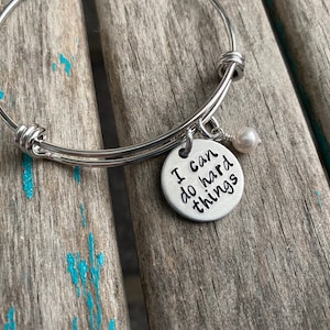 May include: A silver bangle bracelet with a charm that reads "I can do hard things". The charm is attached to a small white pearl.