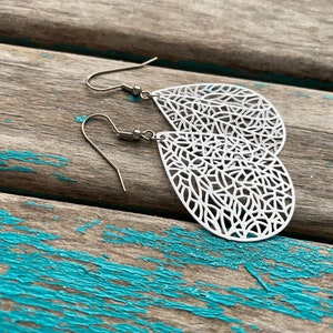May include: A pair of silver teardrop earrings with a delicate, intricate filigree design. The earrings are made of metal and have a hook closure.