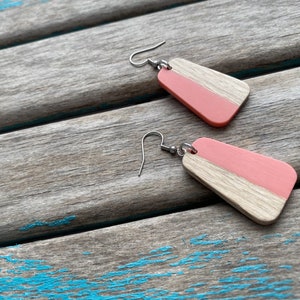 May include: A pair of wooden earrings with a pink and natural wood finish. The earrings are rectangular in shape and have a silver hook.