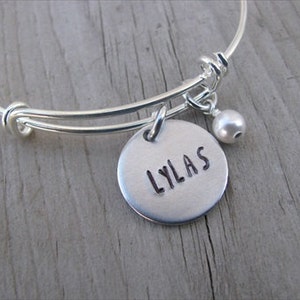 May include: Silver bangle bracelet with a round charm that says "LYLAS" and a small pearl charm.
