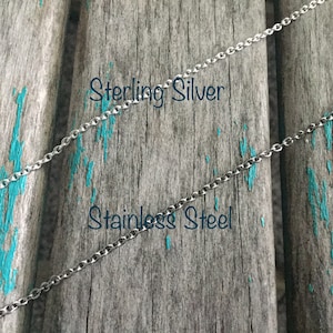 Może przedstawiać: Dwa srebrne łańcuszki na zniszczonym drewnianym tle. G&oacute;rny łańcuszek jest oznaczony jako "Sterling Silver", a dolny jako "Stainless Steel".