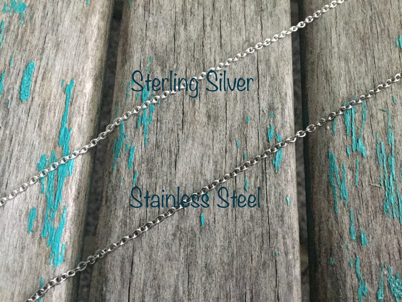 Może przedstawiać: Dwa srebrne łańcuszki na zniszczonym drewnianym tle. G&oacute;rny łańcuszek jest oznaczony jako "Sterling Silver", a dolny jako "Stainless Steel".