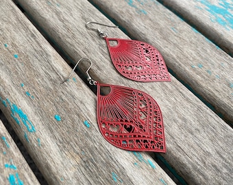 May include: A pair of red filigree leaf-shaped earrings with silver-colored hooks. The earrings have a detailed openwork design with heart and sunburst patterns. The earrings are displayed on a weathered, light blue and gray wooden surface.
