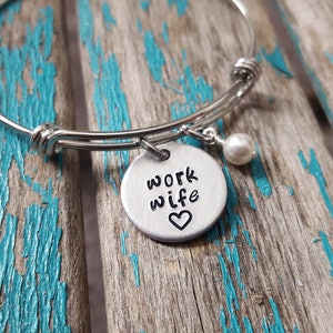 May include: A silver bangle bracelet with a charm that says "work wife" with a small heart. A small white pearl dangles from the charm.