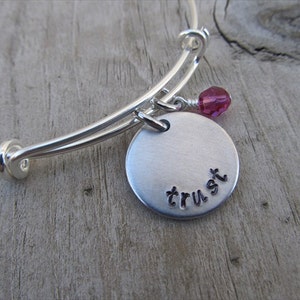 May include: Silver bangle bracelet with a round charm that says "trust" and a pink crystal bead.