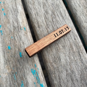 May include: A wooden tie bar with the date 11.07.15 engraved on it. The tie bar is brown and has a natural wood grain.