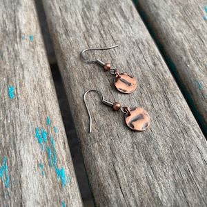 May include: A pair of copper earrings with a hammered texture. The earrings are on a wooden surface.