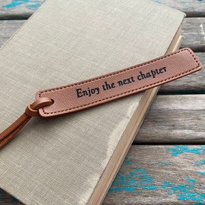 May include: Brown leather bookmark with the text "Enjoy the next chapter" on a book.