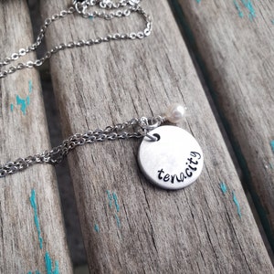 May include: A silver chain necklace with a round silver charm that says "tenacity" and a small white pearl accent.