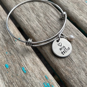 May include: A silver bangle bracelet with a charm that says "my tribe" with a heart above the word "my". A small white pearl dangles from the charm.