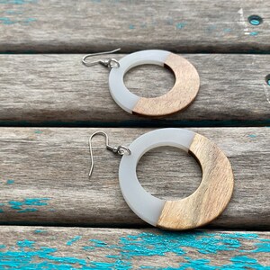 May include: A pair of hoop earrings with a modern design. Each earring features a white, circular section and a section of light-colored wood. The earrings have silver-colored ear wires and are displayed on a weathered wooden surface.