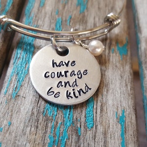 May include: A silver expandable bracelet with a round charm that says "have courage and be kind". The charm has a small pearl attached to it.