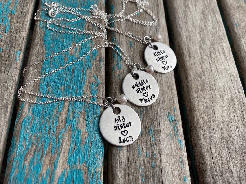 May include: Three silver necklaces with round pendants that say "big sister Lucy", "middle sister Maya", and "little sister Nora". Each pendant has a small pearl charm.