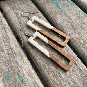 May include: A pair of rectangular dangle earrings. Each earring features a white top section and a brown wooden bottom section. The earrings have silver-colored hooks.