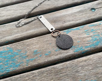 May include: A silver chain necklace with a silver bar pendant and a black circle pendant with a textured surface. The bar pendant has a small silver ring attached to it, and the circle pendant is attached to the ring with a copper ring.
