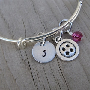 May include: Silver charm bracelet with a letter J charm, a button charm, and a pink crystal charm.