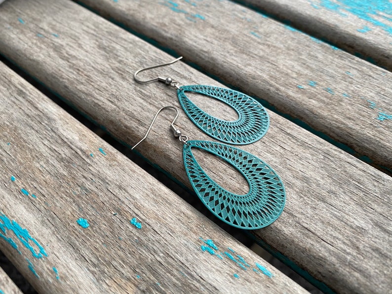 May include: Teal blue teardrop-shaped earrings with a geometric pattern. The earrings are made of metal and have a silver hook.