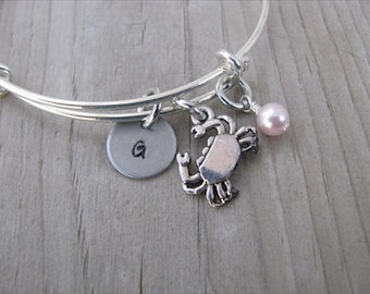 May include: Silver charm bracelet with a crab charm, a pink pearl charm, and a silver disc charm with the letter "G" engraved on it.