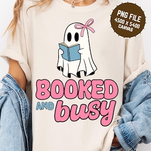 May include: Cream-coloured t-shirt with a cartoon ghost reading a blue book, wearing a pink bow. The text "BOOKED and busy" is in pink and blue. A brown circle with text "PNG FILE 4500 X 5400 CANVAS" is in the upper right corner.