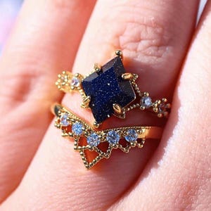 May include: Two gold rings with intricate designs. The top ring features a dark blue, square-cut stone with a sparkling effect. The second ring is a chevron shape with small, clear stones. Both rings are on a finger.