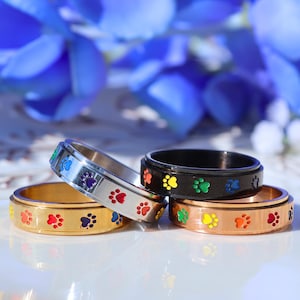 May include: Four colorful spinner rings with paw print designs. The rings are gold, silver, black, and copper, each with a different color paw print. The background features blue and white flowers.