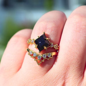 May include: Two gold rings are displayed on a finger. The top ring features a dark blue, star-shaped stone. The second ring is a gold band with a chevron design and small, clear stones. The rings are set against a blurred background.