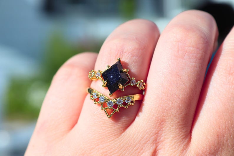 May include: Two gold rings are displayed on a finger. The top ring features a dark blue, star-shaped stone. The second ring is a gold band with a chevron design and small, clear stones. The rings are set against a blurred background.