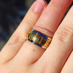 May include: A gold and silver ring with a decorative design featuring red accents. The ring is worn on a finger, showcasing its intricate details and craftsmanship. The ring's design includes gold and red patterns.