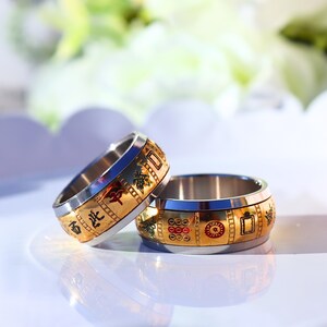 May include: Two stainless steel rings with gold-colored bands. The bands feature decorative patterns and Chinese characters. The rings are on a white surface with a blurred background of flowers.