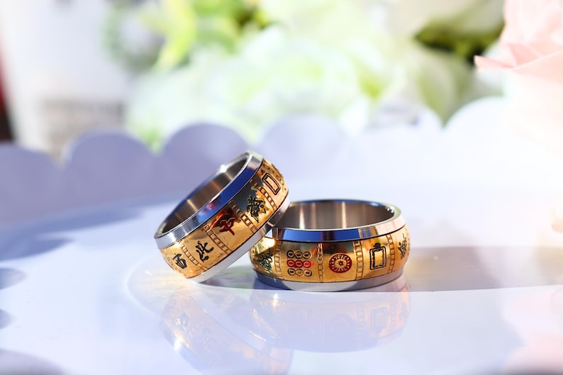 May include: Two stainless steel rings with gold-colored bands. The bands feature decorative patterns and Chinese characters. The rings are on a white surface with a blurred background of flowers.