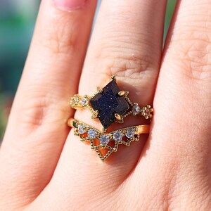 May include: Two gold rings with a dark blue gemstone and clear accent stones. The top ring features a square-cut, dark blue stone. The bottom ring has a V-shaped design with clear stones. The rings are on a finger.