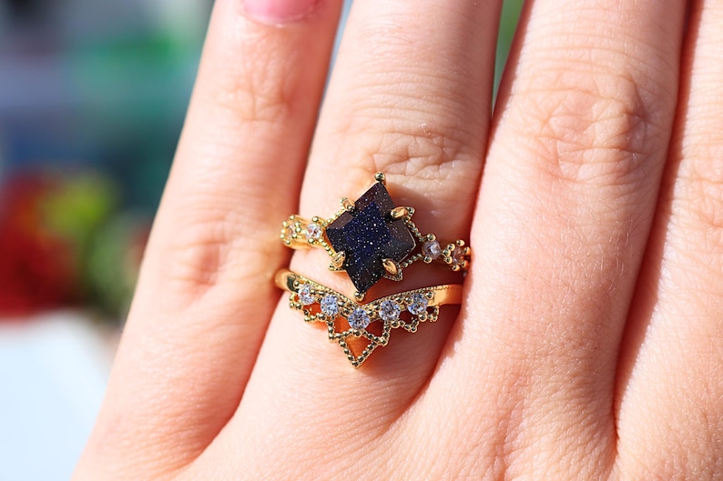 May include: Two gold rings with a dark blue gemstone and clear accent stones. The top ring features a square-cut, dark blue stone. The bottom ring has a V-shaped design with clear stones. The rings are on a finger.