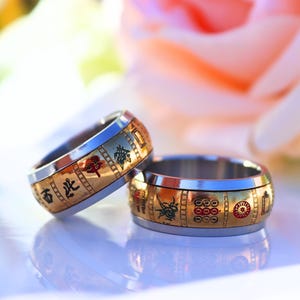 May include: Two wedding rings with a gold band and silver interior. The gold band features Chinese characters and red and green accents. The rings are displayed on a white surface with a blurred pink flower in the background.