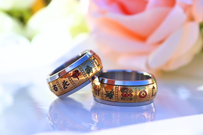 May include: Two wedding rings with a gold band and silver interior. The gold band features Chinese characters and red and green accents. The rings are displayed on a white surface with a blurred pink flower in the background.