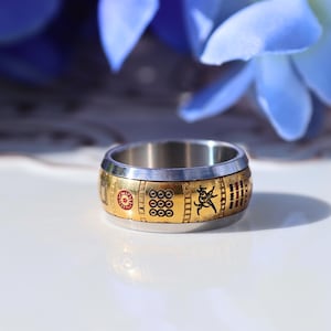 May include: A stainless steel ring with a gold band featuring red and black symbols. The ring has a unique design with a sun, circles, and a figure. The ring is a stylish accessory.