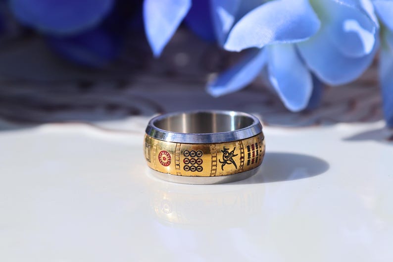 May include: A stainless steel ring with a gold band featuring red and black symbols. The ring has a unique design with a sun, circles, and a figure. The ring is a stylish accessory.
