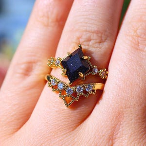 May include: A gold-colored ring set featuring a dark blue, square-cut gemstone as the centerpiece. The ring is accented with smaller, clear gemstones and intricate detailing. A second, chevron-shaped ring with clear gemstones complements the main ring.