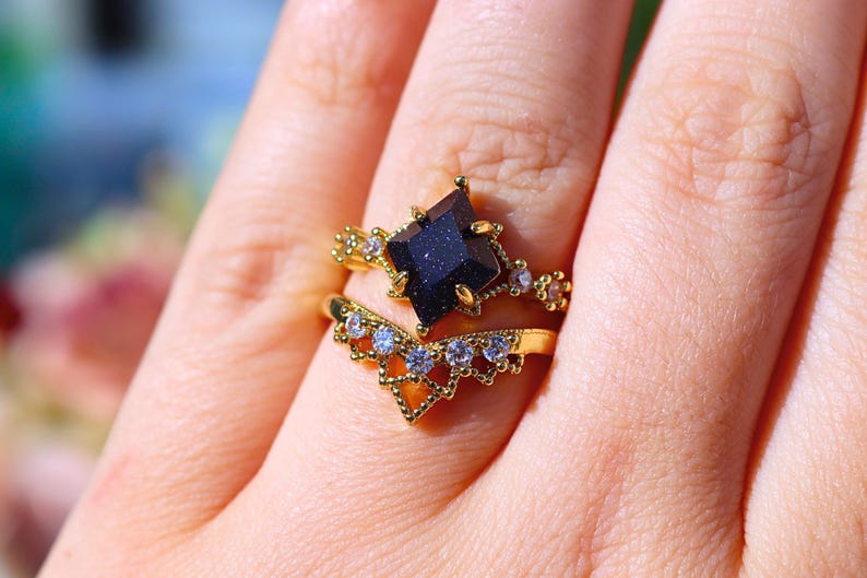 May include: A gold-colored ring set featuring a dark blue, square-cut gemstone as the centerpiece. The ring is accented with smaller, clear gemstones and intricate detailing. A second, chevron-shaped ring with clear gemstones complements the main ring.