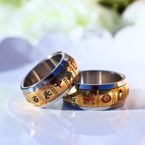 May include: Two gold and silver rings with intricate designs. The rings have a gold band with Chinese characters and symbols, and a silver inner band. The rings are displayed on a white surface with a blurred background.
