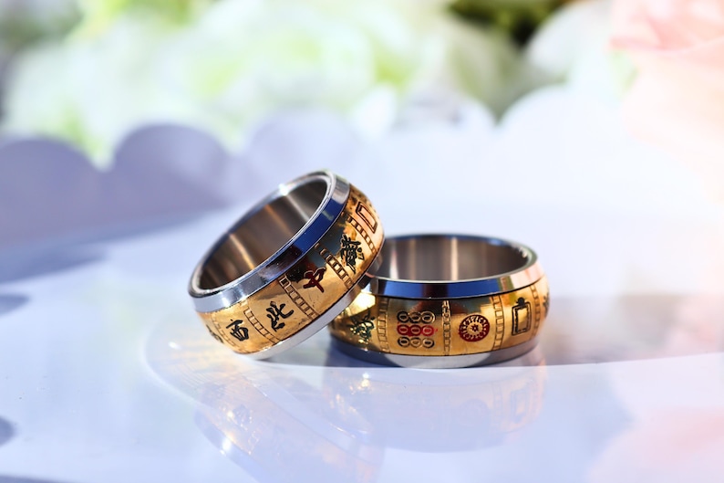 May include: Two gold and silver rings with intricate designs. The rings have a gold band with Chinese characters and symbols, and a silver inner band. The rings are displayed on a white surface with a blurred background.