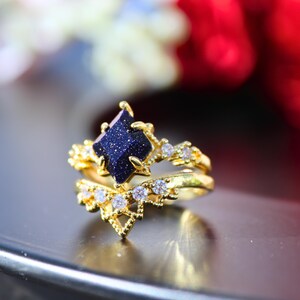 May include: A gold-colored ring set featuring a dark blue gemstone with a sparkling appearance. The set includes a main ring with a square-shaped stone and a matching band, both adorned with small, clear gemstones.