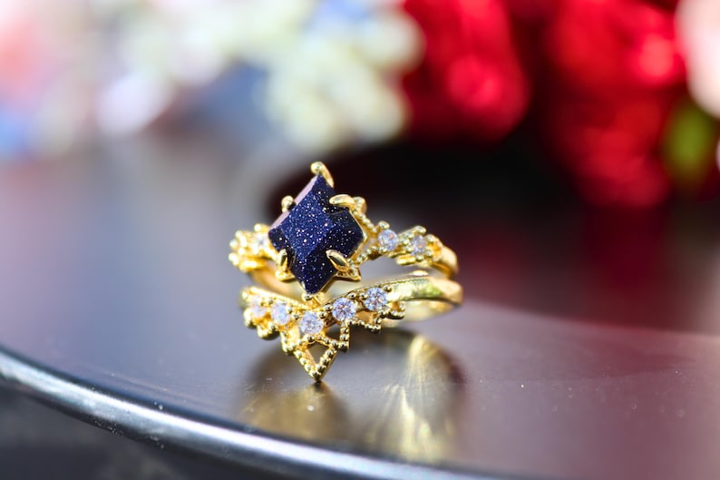 May include: A gold-colored ring set featuring a dark blue gemstone with a sparkling appearance. The set includes a main ring with a square-shaped stone and a matching band, both adorned with small, clear gemstones.