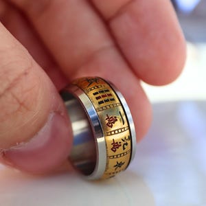 May include: A two-tone ring with a gold-colored band featuring engraved characters and a silver-colored inner band. The ring is held between fingers, showcasing its design and craftsmanship. The ring's design includes black characters.