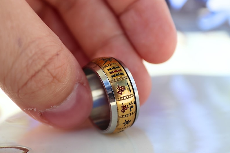 May include: A two-tone ring with a gold-colored band featuring engraved characters and a silver-colored inner band. The ring is held between fingers, showcasing its design and craftsmanship. The ring's design includes black characters.