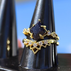 May include: A gold-colored ring set. The engagement ring features a dark blue, geometric-shaped stone with gold accents and small clear stones. The wedding band is a gold band with a V-shaped design and small clear stones.