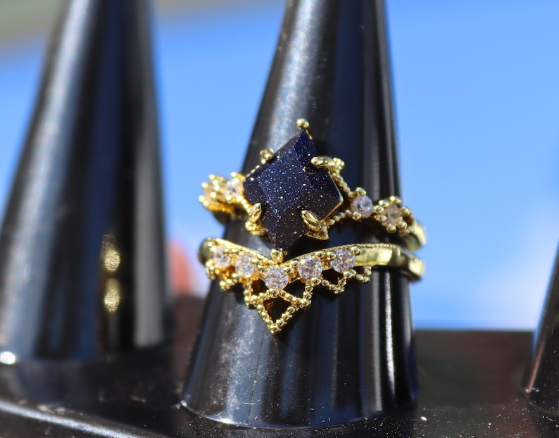 May include: A gold-colored ring set. The engagement ring features a dark blue, geometric-shaped stone with gold accents and small clear stones. The wedding band is a gold band with a V-shaped design and small clear stones.