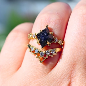 May include: Two gold rings are displayed on a finger. The top ring features a dark blue, square-cut gemstone. The second ring is a gold chevron band with small, clear gemstones. The rings are set against a blurred background.