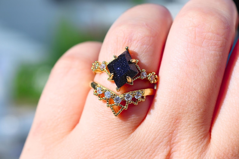 May include: Two gold rings are displayed on a finger. The top ring features a dark blue, square-cut gemstone. The second ring is a gold chevron band with small, clear gemstones. The rings are set against a blurred background.