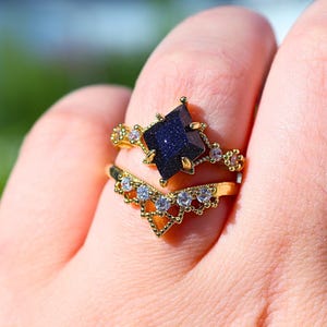 May include: Two gold rings are displayed on a finger. The top ring features a dark blue, square-cut gemstone. The second ring is a chevron shape with small, clear gemstones. Both rings have intricate gold detailing.